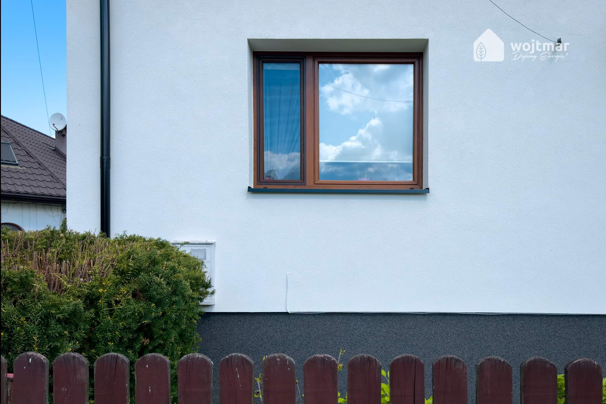 Fragment ocieplonej elewacji domu z brązowym oknem i widocznym logo firmy Wojtmar Dajemy Energię! w prawym górnym rogu.
