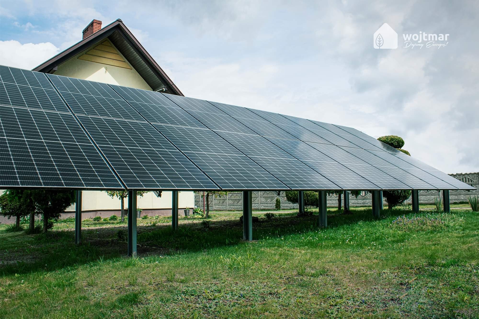 Instalacja fotowoltaiczna na metalowych podporach, ustawiona na trawniku przed domem jednorodzinnym, widoczny fragment ogrodzenia i logo firmy w prawym górnym rogu.