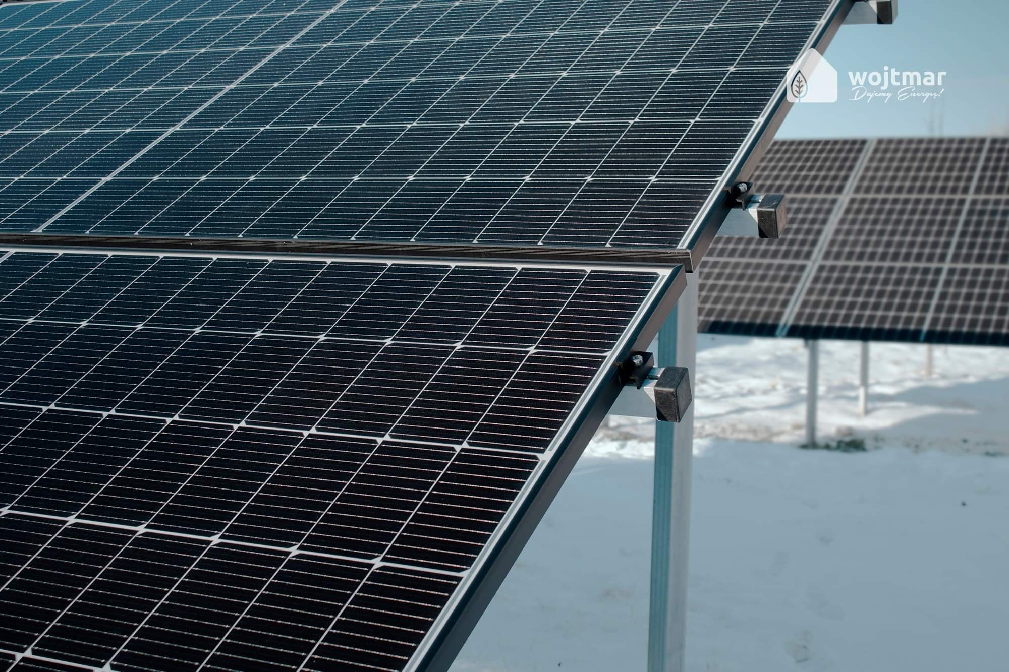 Dwa rzędy paneli fotowoltaicznych na metalowych stelażach, częściowo pokryte śniegiem, z logo firmy 'wojtmar' w prawym górnym rogu.