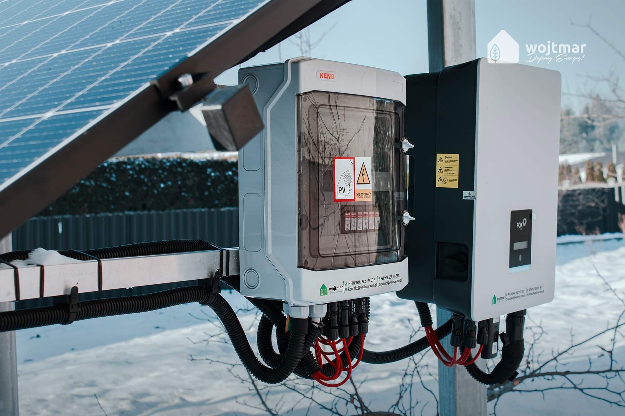 Instalacja fotowoltaiczna w zimowym krajobrazie: skrzynka KENO, inwerter FOX, okablowanie w czarnych peszlach, panele słoneczne w tle pokryte śniegiem.