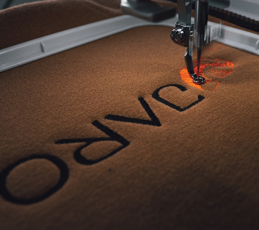 Proces haftowania ciemnych liter na brązowym materiale przy użyciu maszyny do haftowania; igła maszyny w trakcie pracy nad literą 'C'.