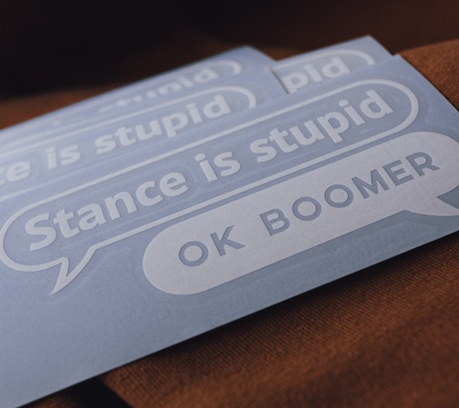 Stos naklejek z napisem 'Stance is stupid' i 'OK BOOMER' w dymkach dialogowych, leżących na brązowej tkaninie.