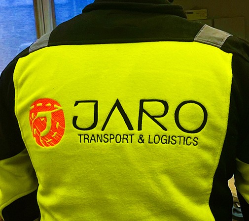 Żółta bluza robocza z czarnymi ramionami, na plecach wyhaftowane logo firmy Jaro Transport & Logistics, z lewej strony logo w kształcie litery J z pomarańczowym wypełnieniem.
