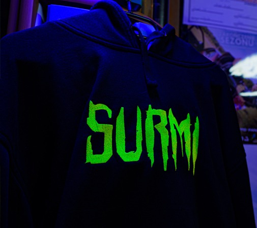 Czarna bluza z kapturem z jaskrawozielonym napisem 'SURMI' w stylu gotyckim, oświetlona niebieskim światłem.