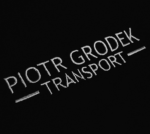 Czarna tkanina z wyhaftowanym białym napisem 'PIOTR GRODEK TRANSPORT' z podkreśleniami po bokach, wykonanym w technice haftu płaskiego.