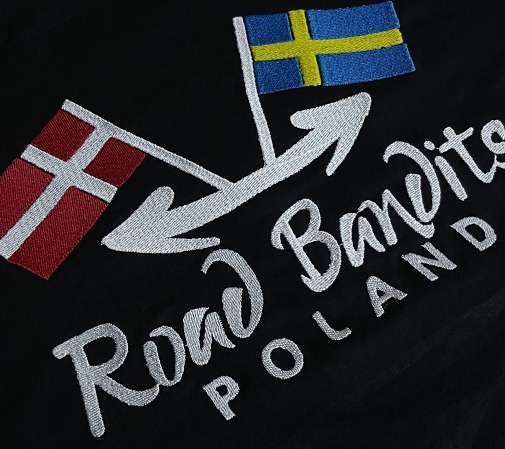 Czarna tkanina z haftowanym napisem 'Road Bandits POLAND' oraz flagami Danii i Szwecji połączonymi strzałkami, wykonane srebrną nicią.