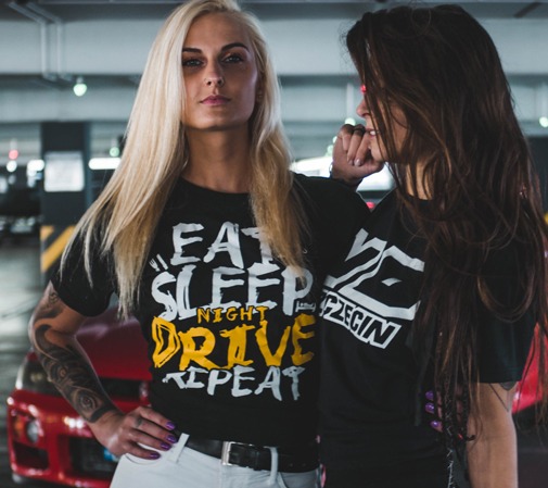Dwie kobiety pozują w czarnych koszulkach z nadrukami na tle parkingu podziemnego i czerwonego samochodu. Kobieta z blond włosami ma tatuaż na ramieniu i koszulkę z napisem 'EAT SLEEP NIGHT DRIVE...
