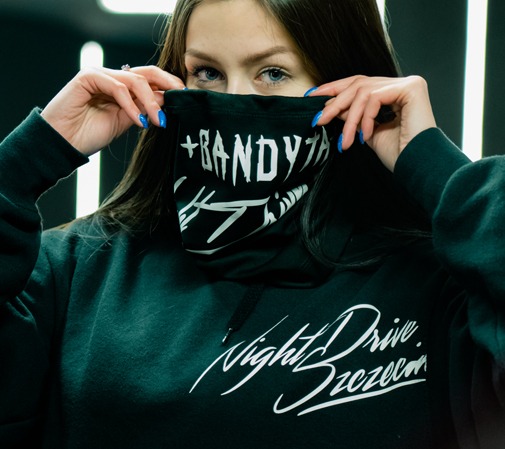 Młoda kobieta w czarnej bluzie z kapturem z białym napisem 'Night Drive Szczecin' zasłaniająca twarz czarną chustą z białym napisem '+BANDYTHA #SZCZECIN', widoczne niebieskie paznokcie.