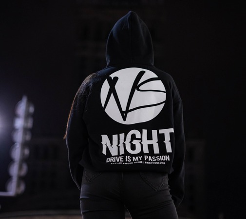 Osoba w czarnej bluzie z kapturem, na której plecach widnieje biały okrągły nadruk z abstrakcyjnym wzorem, poniżej napis 'NIGHT' z efektem rozmycia, a jeszcze niżej slogan 'DRIVE IS MY PASSION'...