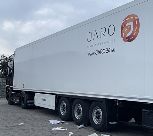 Biała naczepa ciężarówki z dużym logo firmy transportowej JARO, widoczne koła i fragment kabiny, na ziemi leżą resztki folii po naklejeniu logo.