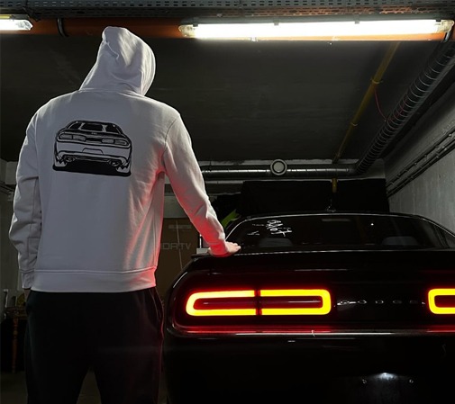 Osoba w białej bluzie z kapturem, na której plecach widnieje czarny nadruk przedstawiający tył samochodu, stoi obok czarnego samochodu z włączonymi tylnymi światłami. Scena w garażu.