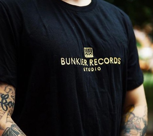 Czarna koszulka z haftowanym złotym logo 'Bunkier Records Studio', widoczne tatuaże na ramieniu.