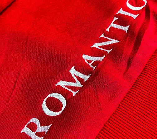 Czerwona bluza z białym haftowanym napisem 'ROMANTIC' biegnącym po skosie, widoczny fragment ściągacza.