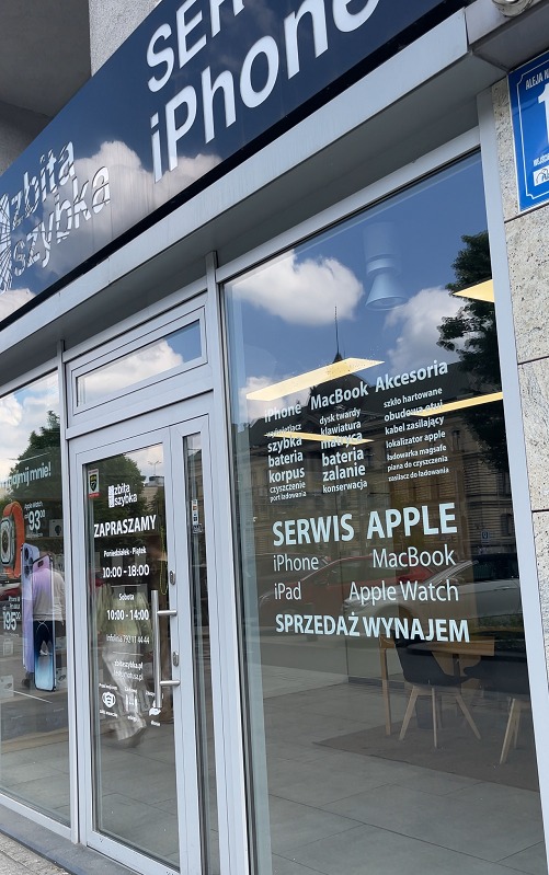 Wejście do serwisu Apple z widocznym szyldem 'SERWIS iPhone' oraz informacjami o naprawach i sprzedaży akcesoriów. Na szybie naklejone godziny otwarcia i lista usług.