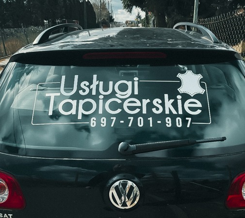 Czarny samochód z naklejką na tylnej szybie reklamującą usługi tapicerskie i numer telefonu, widok z tyłu.