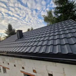 Nowe, czarne pokrycie dachowe z blachodachówki modułowej, zamontowane na budynku w trakcie budowy, widoczne rynny, komin i błękitne niebo z chmurami.