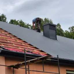 Montaż blachy falistej na dachu domu jednorodzinnego. Widoczny fragment więźby dachowej i komin. Prace na wysokości zabezpieczone rusztowaniem i drabiną.