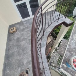 RAF-STONE Rafał Ćwierz - Zewnętrzne, kręcone schody z brązową balustradą na szarym tarasie z płyt. Widoczna drabina, wiadro z farbą i fragment sylwetki osoby. Tło z zielenią drzew.