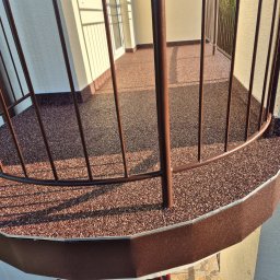 RAF-STONE Rafał Ćwierz - Wykończony balkon z brązową, żywiczną posadzką z kamyczków i metalową balustradą, widok z góry, z delikatnym cieniem słonecznym na powierzchni.
