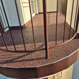 Wykończony balkon z brązową, żywiczną posadzką z kamyczków i metalową balustradą, widok z góry, z delikatnym cieniem słonecznym na powierzchni.