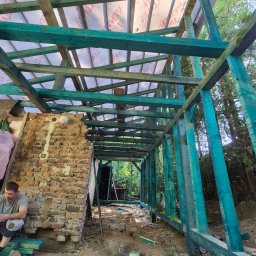 RAF-STONE Rafał Ćwierz - Wnętrze budowy domu: zielona drewniana konstrukcja więźby dachowej z widoczną membraną dachową i fragmentem ceglanej ściany. Mężczyzna siedzi obok ściany.