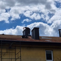 RAF-STONE Rafał Ćwierz - Praca na dachu: dekarz pochylony nad kominem na tle błękitnego nieba z obłokami. Widoczny fragment dachu z deskami i rusztowanie przy budynku.