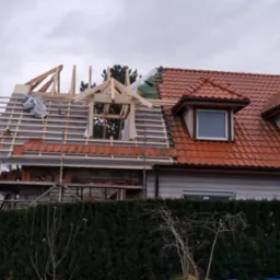 Dom w trakcie wymiany dachu: widoczna więźba i częściowo ułożona dachówka, kontrastujące z istniejącym, czerwonym dachem. Prace na wysokości, rusztowania.