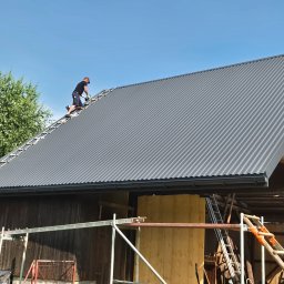 Cezar usługi budowlane - Dekarz w czarnym stroju na drabinie dachowej, montujący ciemnoszarą blachę trapezową na dachu drewnianego budynku, z widoczną rynną i błękitnym niebem.