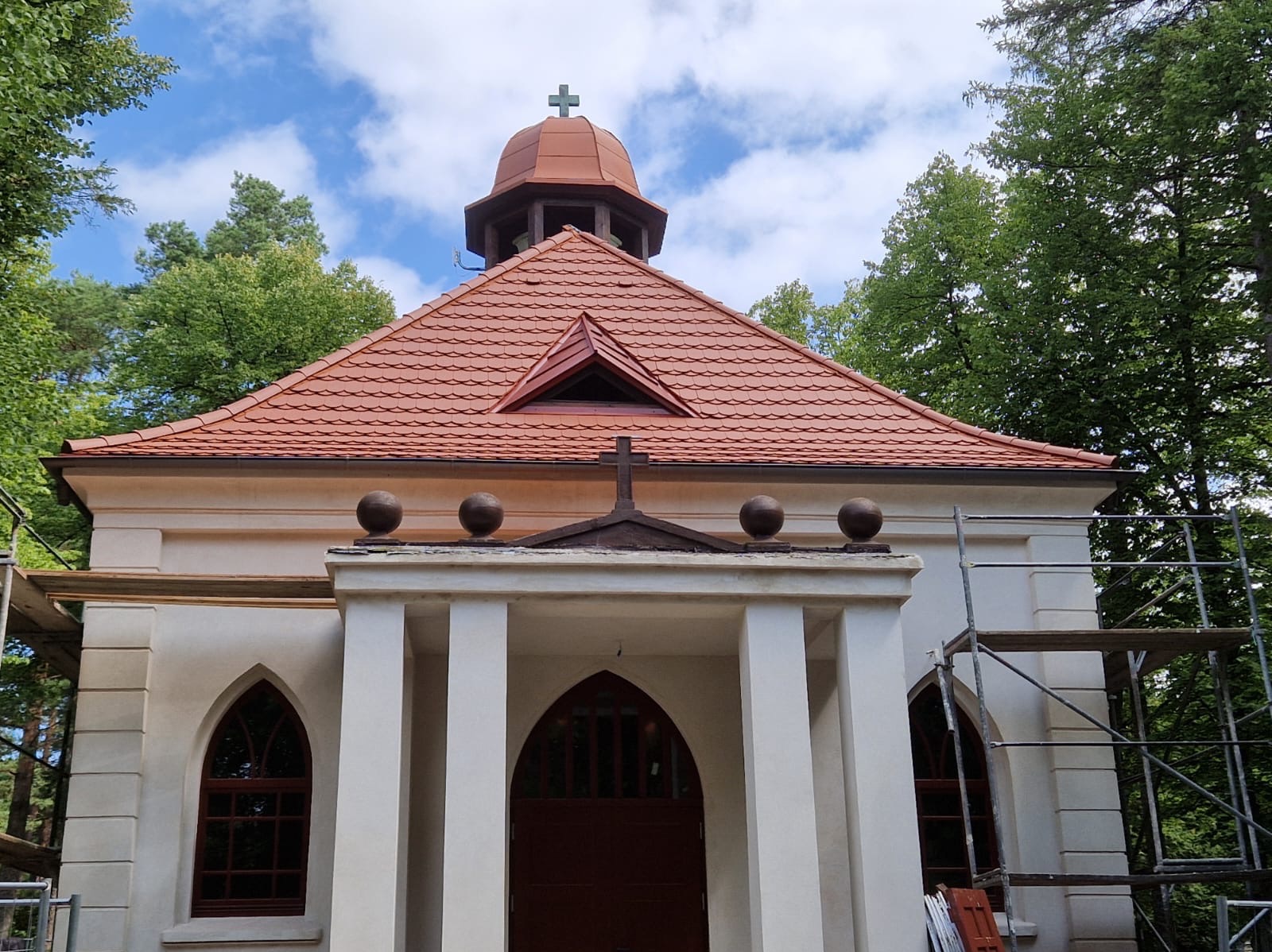 Odnowiona kapliczka z nową, czerwoną dachówką karpiówką i obróbkami blacharskimi, widoczna z dołu na tle błękitnego nieba z chmurami, z fragmentem rusztowania po bokach.