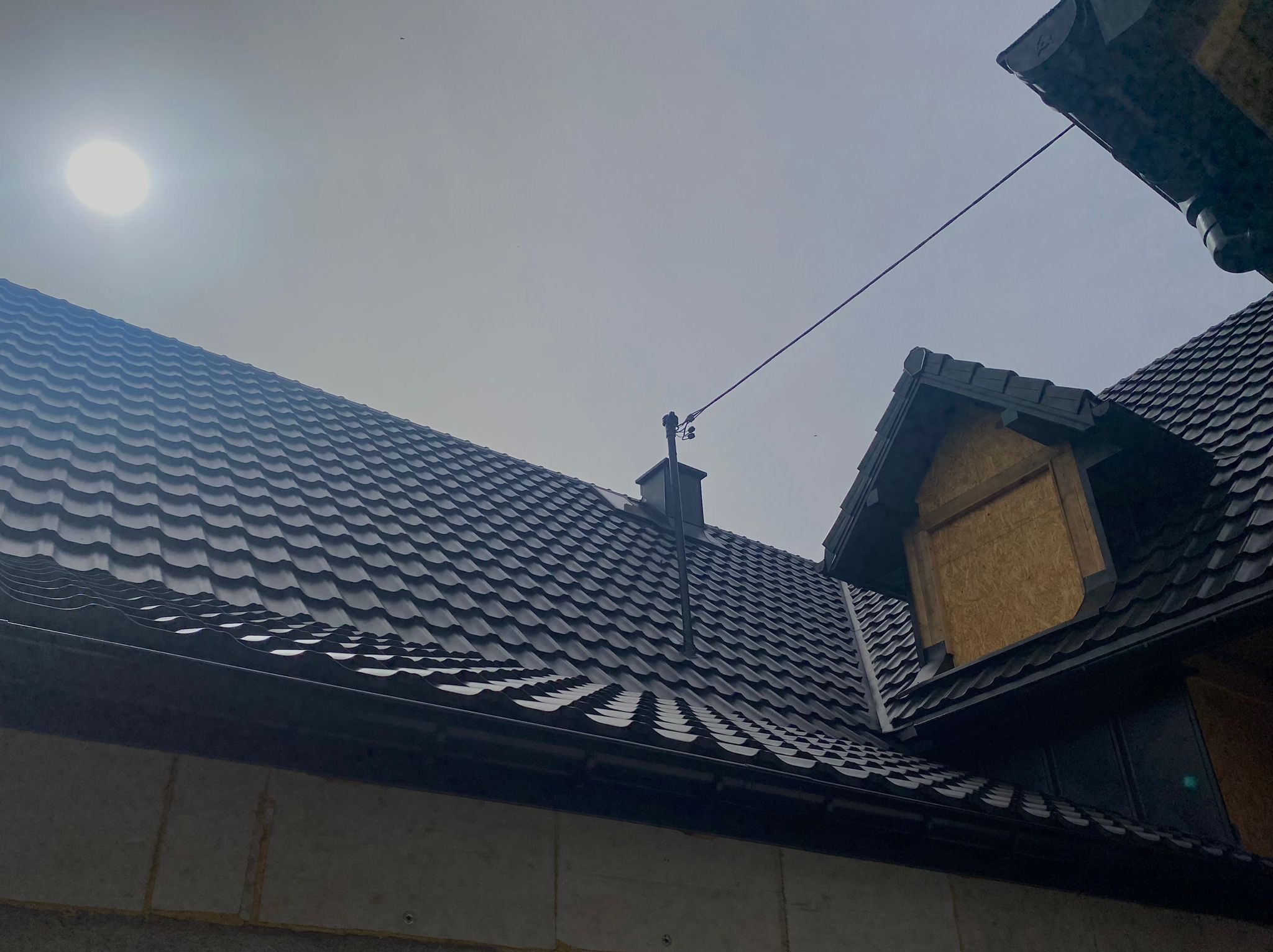 Fragment dachu z czarnej blachodachówki falistej, widoczne rynny, komin i okno dachowe zabite płytą OSB pod pochmurnym niebem.