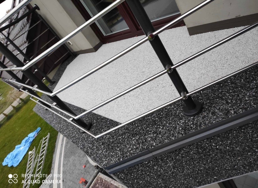 Barierka balkonowa ze stali nierdzewnej z czarnymi słupkami na balkonie wykończonym szarym kamieniem, widok z góry, drabina i plandeka w tle.