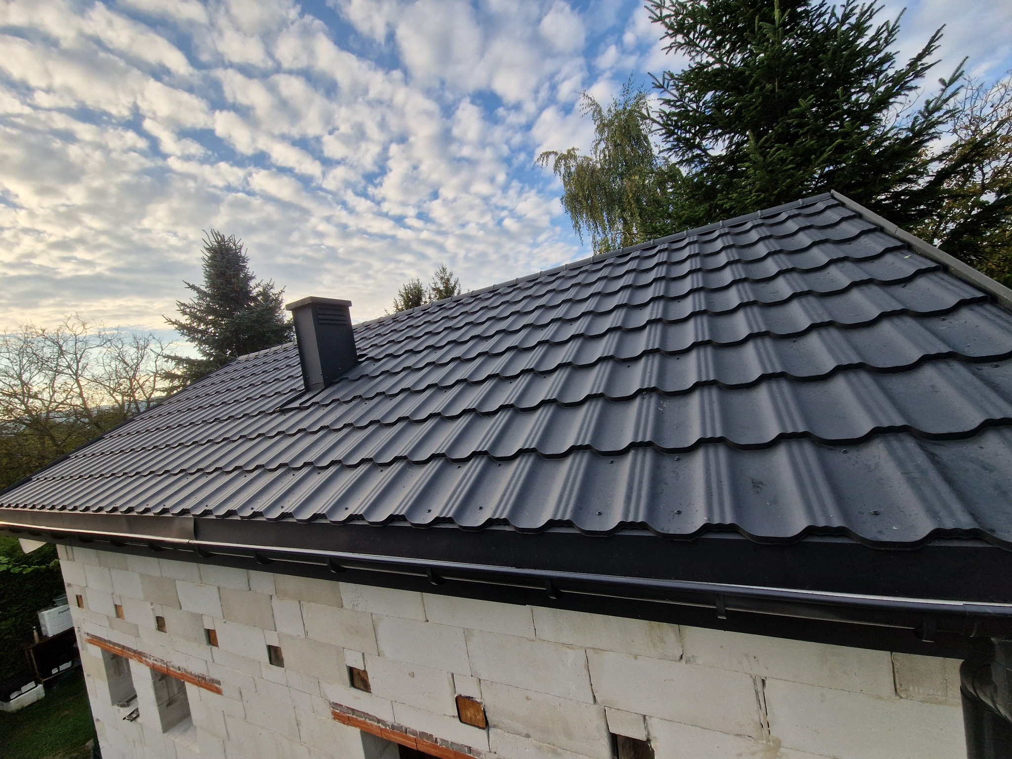 Nowe, czarne pokrycie dachowe z blachodachówki modułowej, zamontowane na budynku w trakcie budowy, widoczne rynny, komin i błękitne niebo z chmurami.