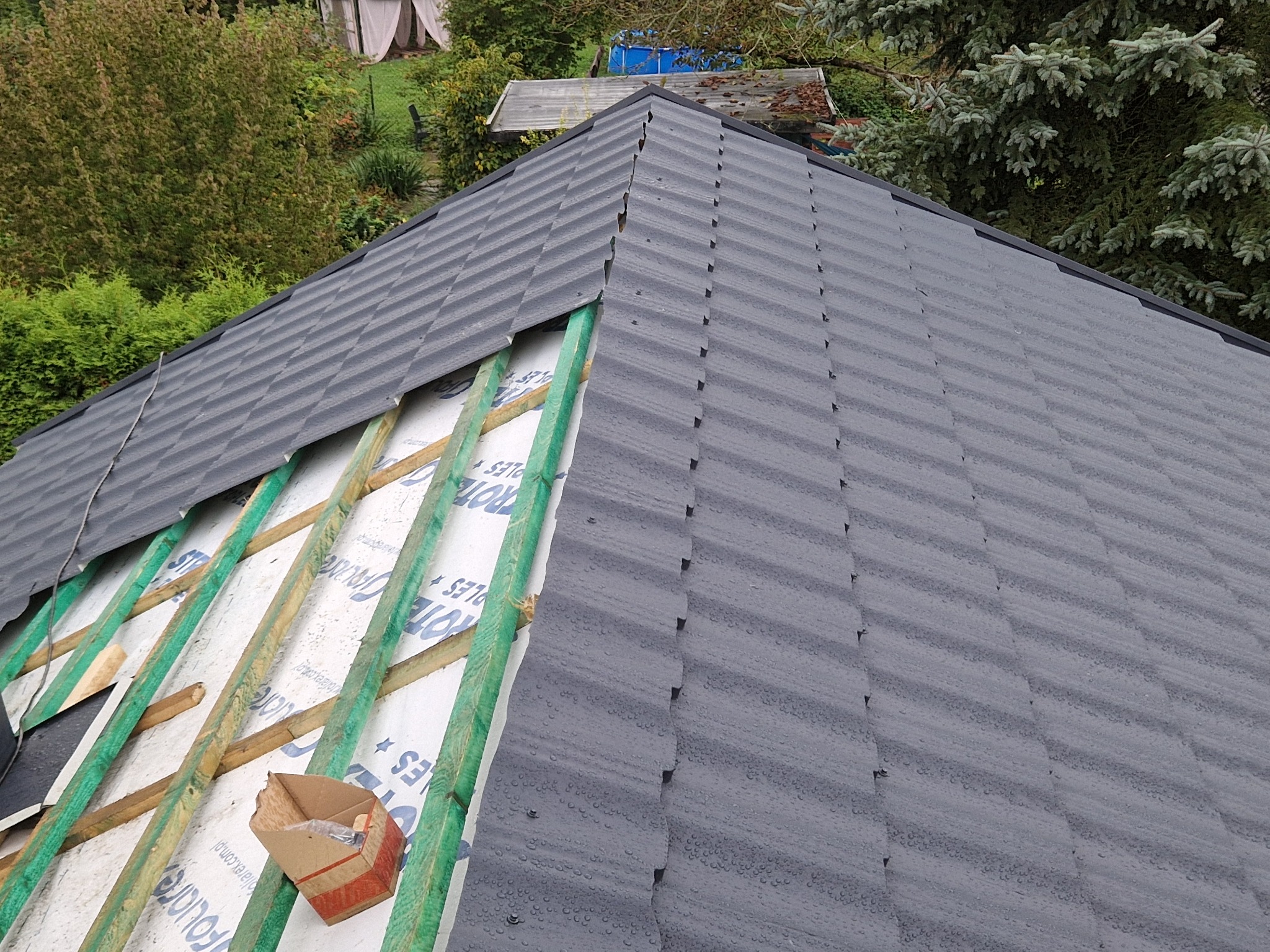 Fragment dachu w trakcie montażu, widoczne krokwie, membrana dachowa i szara blachodachówka z kroplami wody. W tle zieleń drzew i fragment zabudowań.