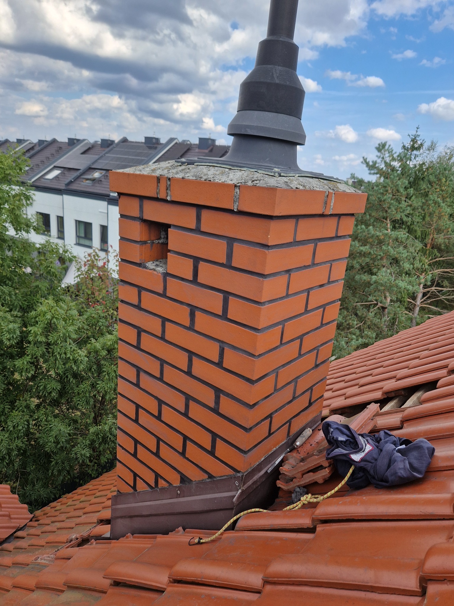 Ceglany komin z czarnym daszkiem na pomarańczowym dachu z dachówki ceramicznej; widoczna lina, ubranie i narzędzie dekarskie, w tle domy i drzewa.