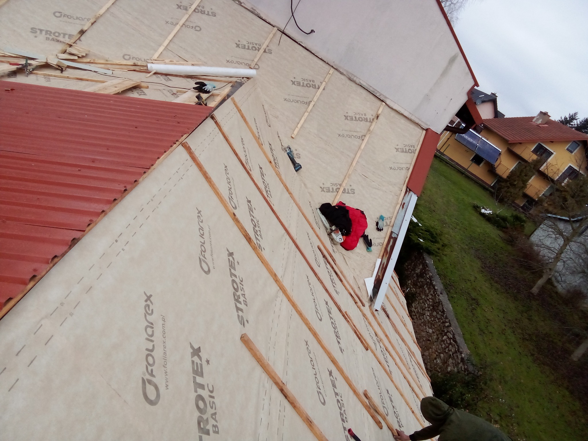 Prace na dachu: montaż folii paroizolacyjnej z widocznymi łatami i częściowo ułożonym czerwonym pokryciem dachowym, pracownik w czerwonej kurtce i czarnych spodniach, narzędzia pracy leżą na dachu.