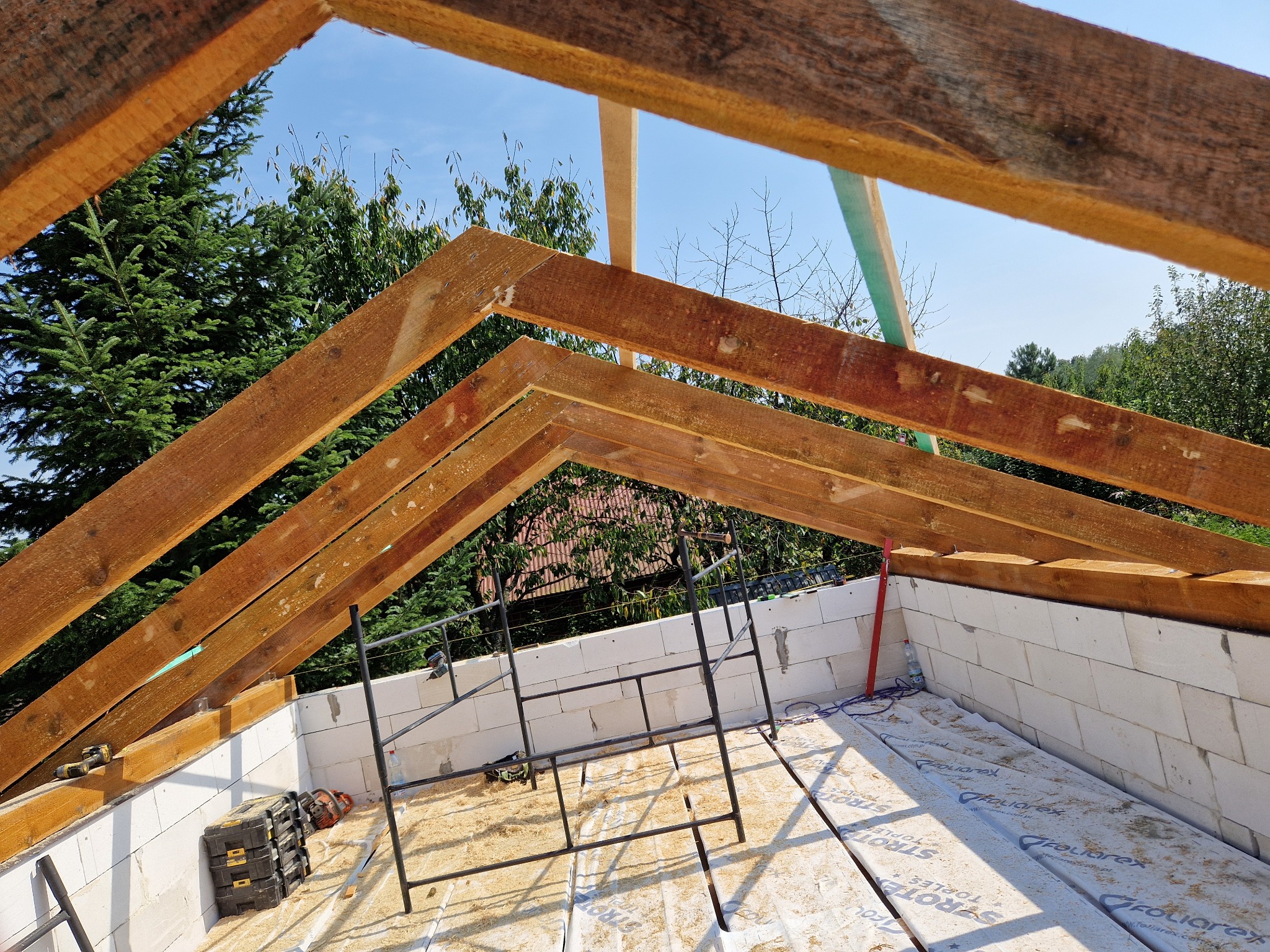 Wznoszenie drewnianej więźby dachowej na budynku z białych bloczków, widoczne rusztowanie, skrzynka narzędziowa i zielone drzewa w tle.