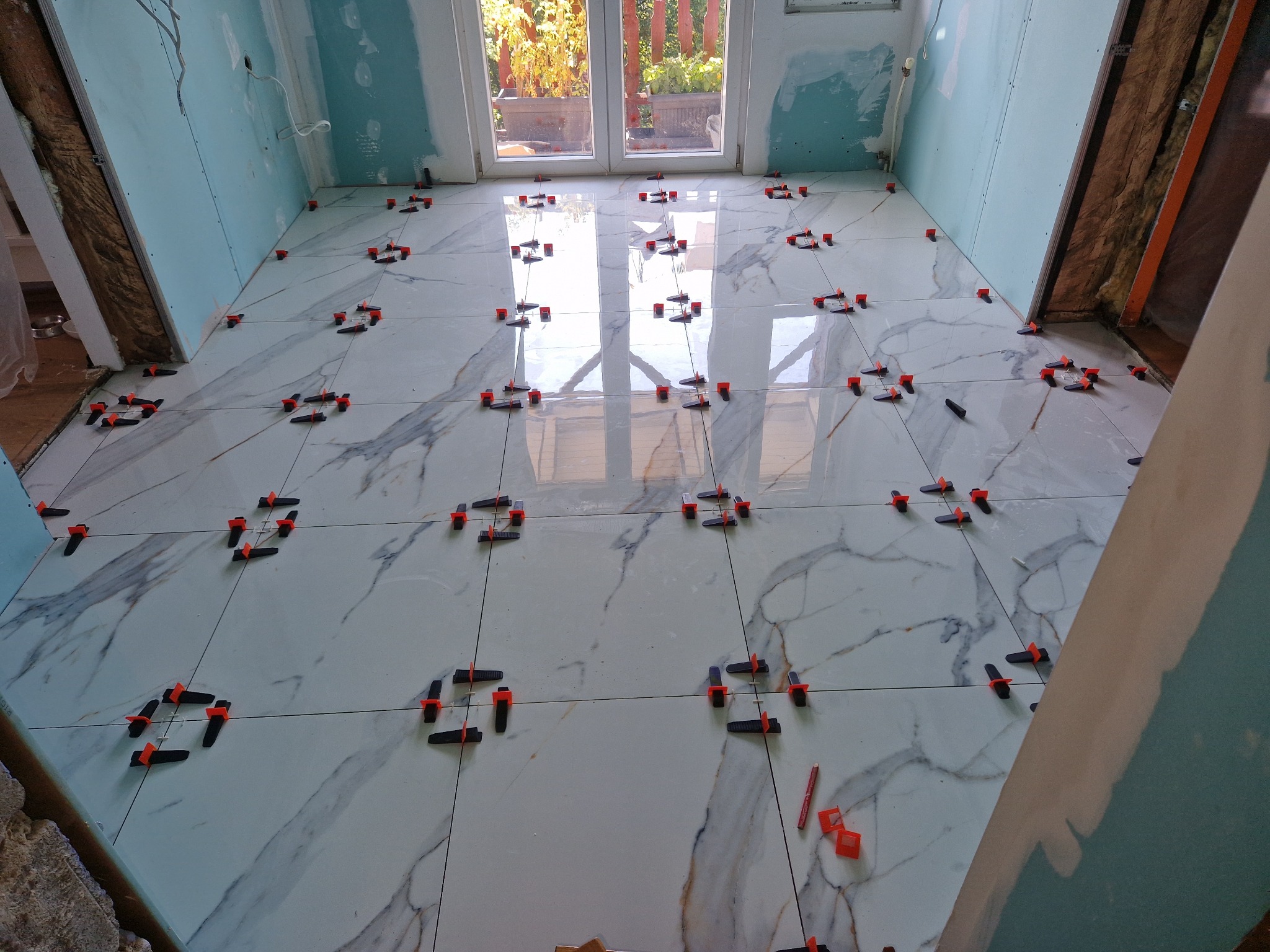 Układanie płytek podłogowych w jasnym wnętrzu z widocznymi klipsami poziomującymi i oknem balkonowym w tle.