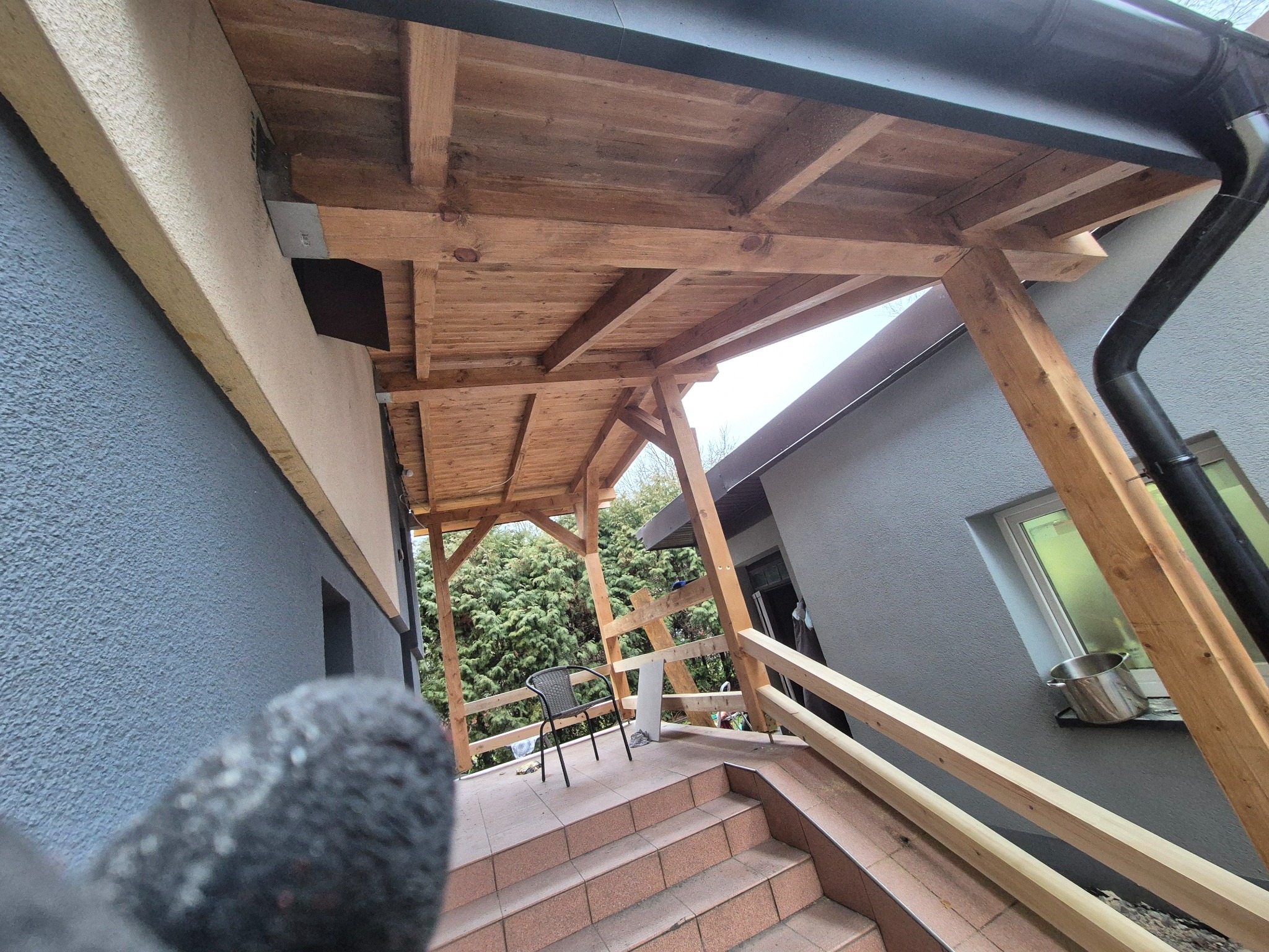 Drewniana konstrukcja zadaszenia tarasu z widoczną więźbą, rynnami i schodami wyłożonymi płytkami. W tle zieleń drzew i krzewów.