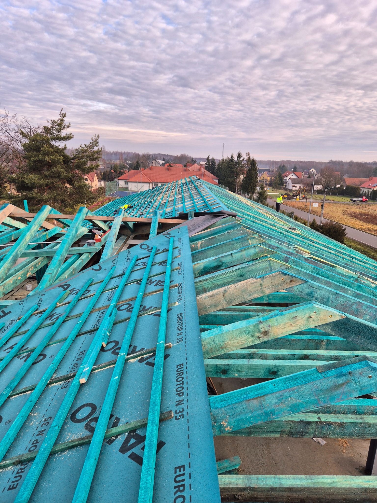 Wysoki kąt na konstrukcję dachu z zielonymi belkami i membraną EUROTOP N-35, w tle zabudowa jednorodzinna pod pochmurnym niebem.
