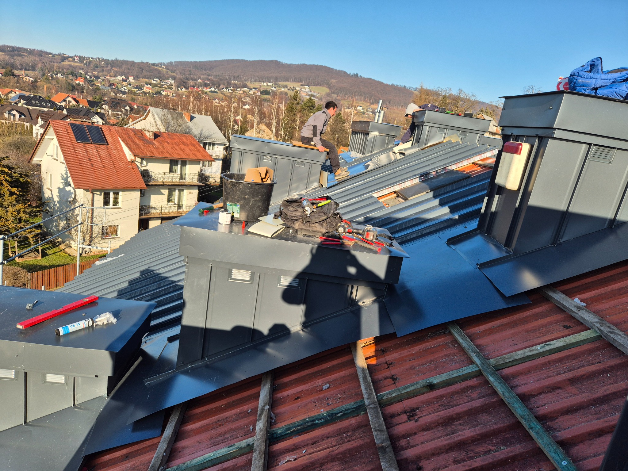 Dekarze montują blaszane elementy na dachu domu w słoneczny dzień. Widoczne narzędzia, poziomica i panorama miasta w tle. Dach pokryty czerwoną dachówką.