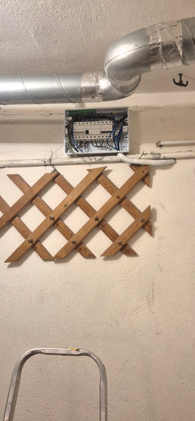 Otwarta skrzynka elektryczna z przewodami na ścianie, poniżej drewniana krata dekoracyjna. Widoczny fragment drabiny i rura wentylacyjna pod sufitem.