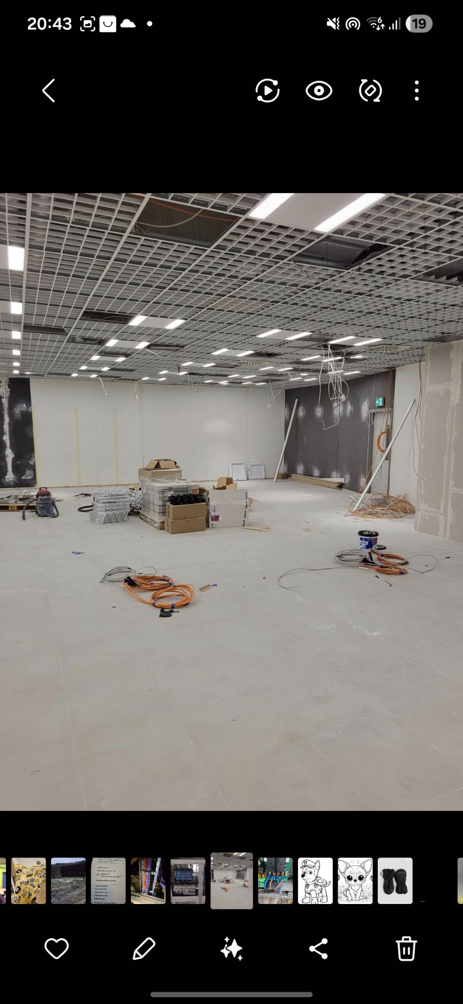 Wnętrze w trakcie prac wykończeniowych: podwieszany sufit z oświetleniem, kable, materiały budowlane. Widok na przestronne pomieszczenie w trakcie instalacji.