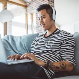 Młody mężczyzna w słuchawkach, w paski, siedzi na szarej sofie z laptopem, z tatuażami na ramieniu, uczący się lub pracujący zdalnie w jasnym salonie.