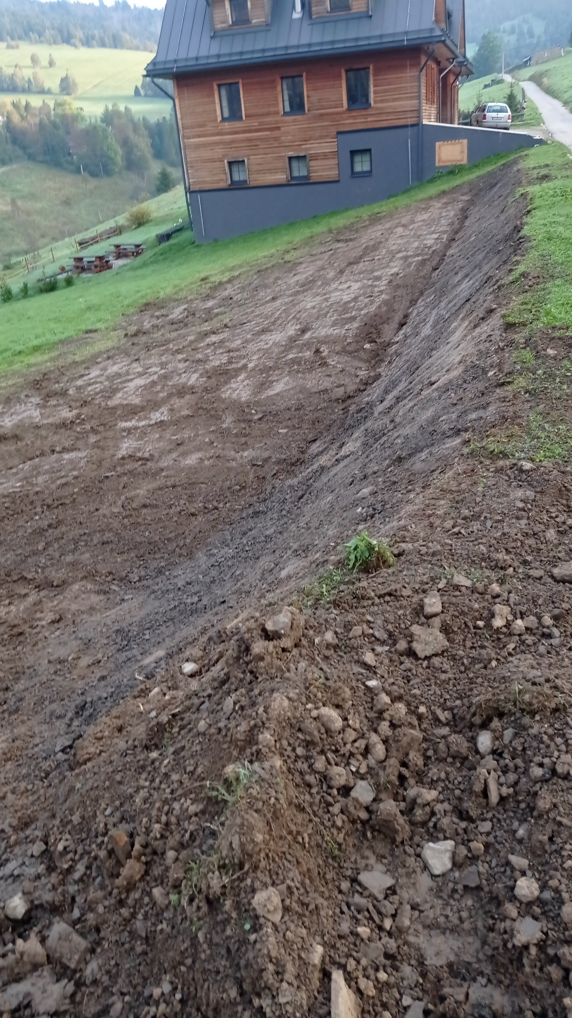 Przygotowany teren pod budowę domu na zboczu, widoczne ślady po maszynach, świeżo wyrównana ziemia i pryzma ziemi z kamieniami na pierwszym planie.