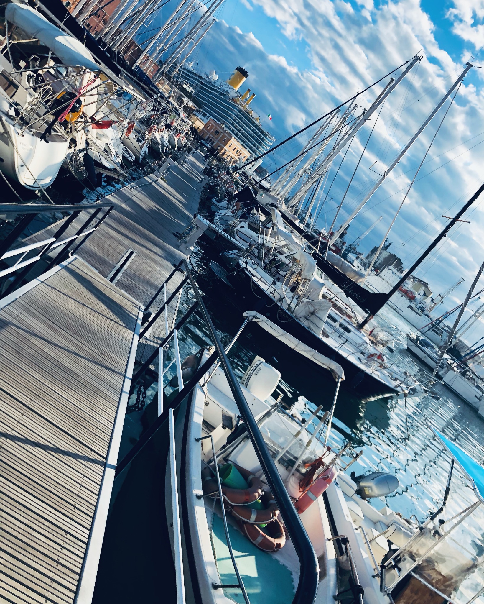 Widok z góry na przystań pełną jachtów żaglowych i motorowych, z drewnianym pomostem i dużym statkiem wycieczkowym w tle pod błękitnym niebem z chmurami.