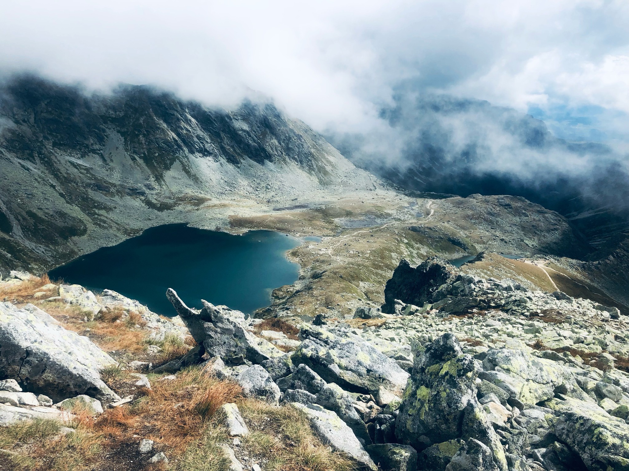 Panoramiczny widok z góry na ciemne jezioro otoczone skalistymi szczytami górskimi spowitymi mgłą i chmurami.
