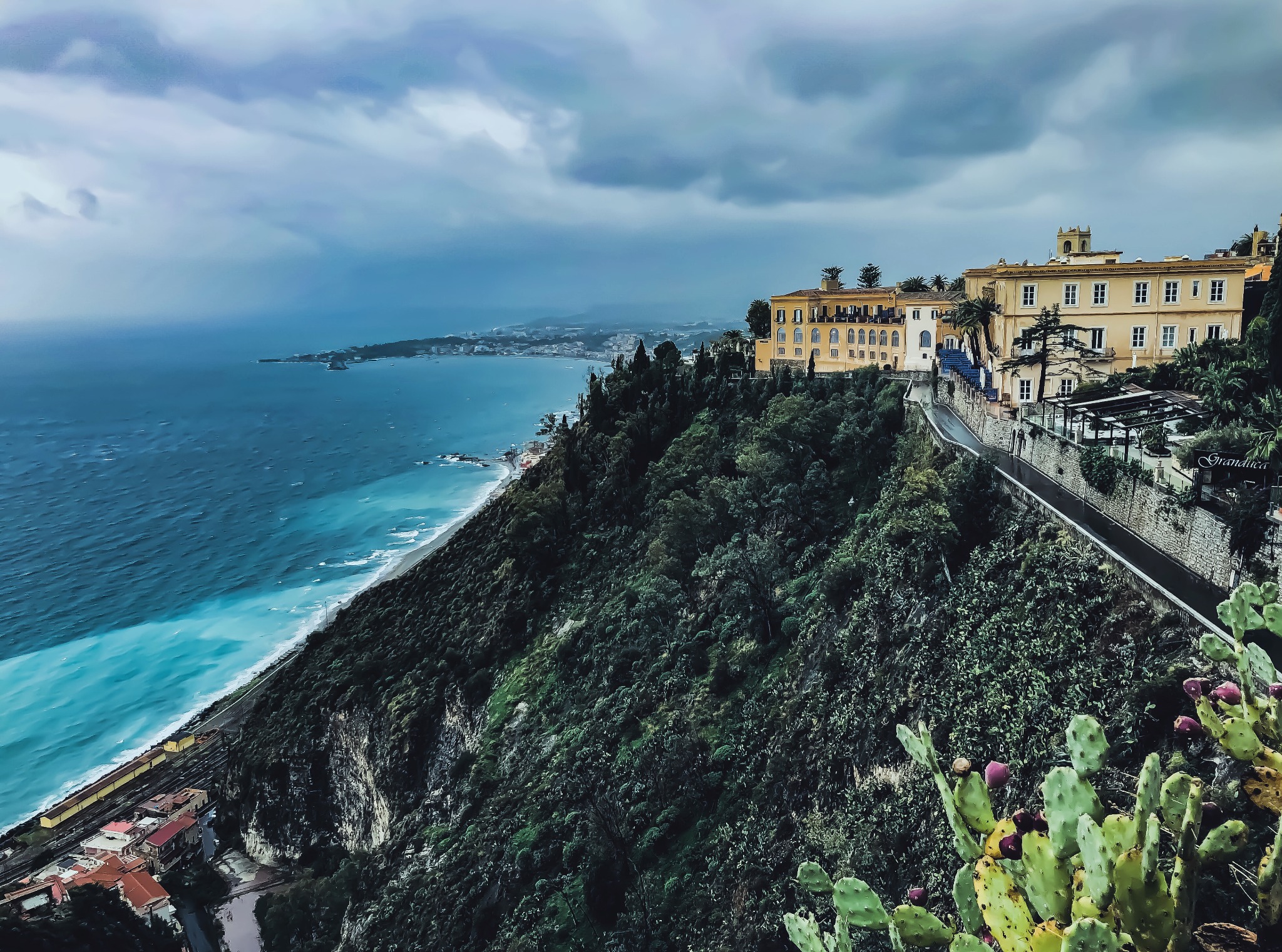 Panoramiczny widok na nadmorski krajobraz z budynkami na wzgórzu, błękitne morze i kaktusy na pierwszym planie.