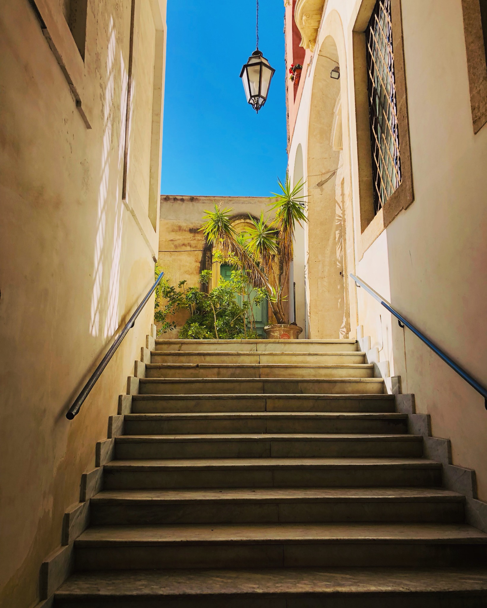 Marmurowe schody prowadzące do zielonych drzwi z roślinami, oświetlone wiszącą latarnią na tle błękitnego nieba.