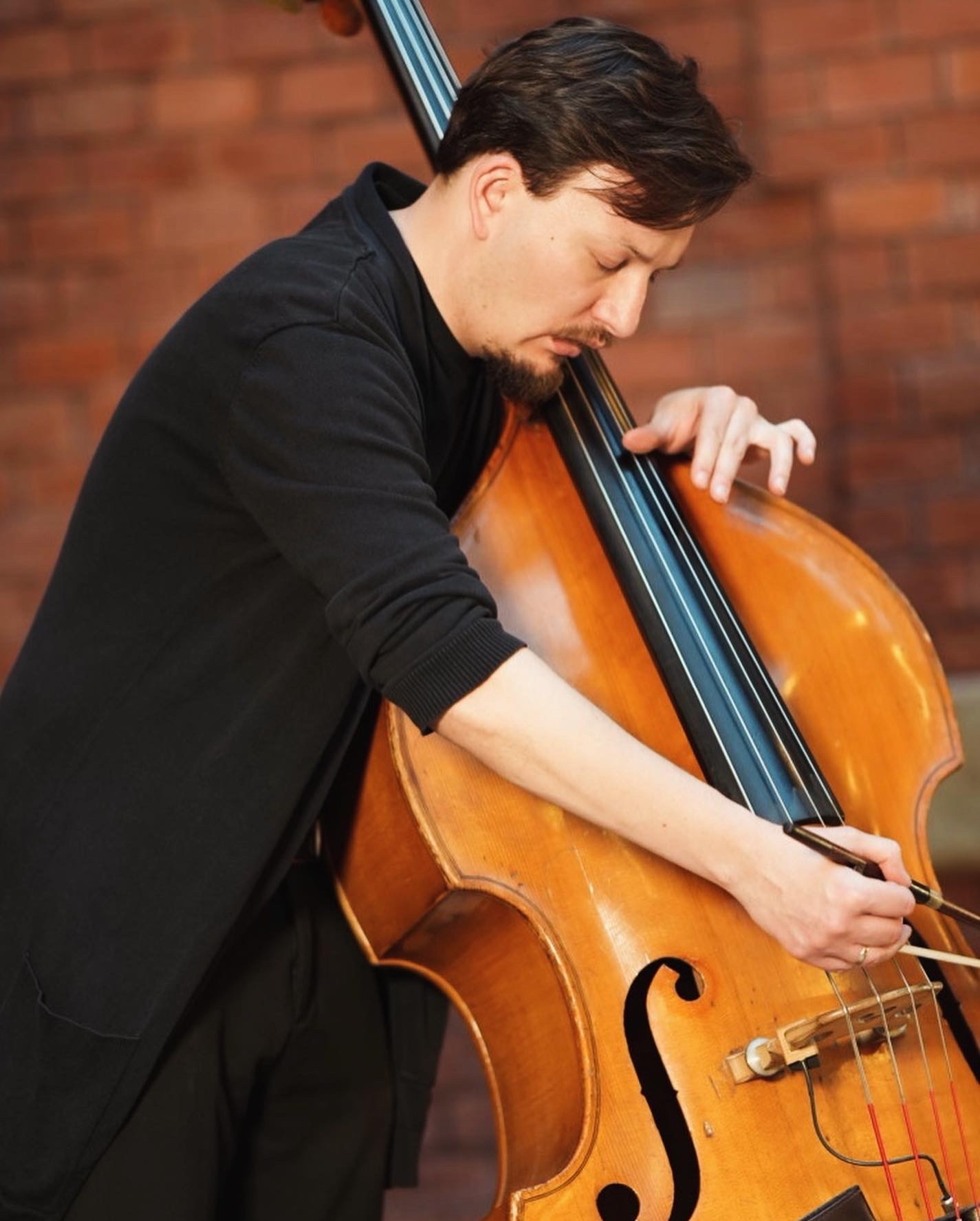 Mężczyzna w czarnym swetrze grający na kontrabasie na tle ceglanej ściany. Skupiony muzyk z brodą trzyma smyczek w prawej dłoni.