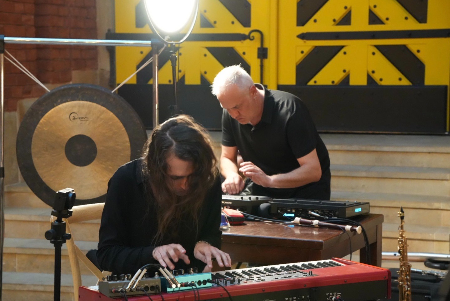 Dwaj muzycy przygotowują się do występu na zewnątrz, z gongiem i oświetleniem studyjnym w tle, klawiszowiec gra na czerwonym keyboardzie, drugi muzyk ustawia sprzęt.